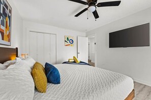 Ein Schlafzimmer mit einem Bett, einem Deckenventilator, einem an der Wand montierten Fernseher und einer Tür zu einem weiteren Raum.