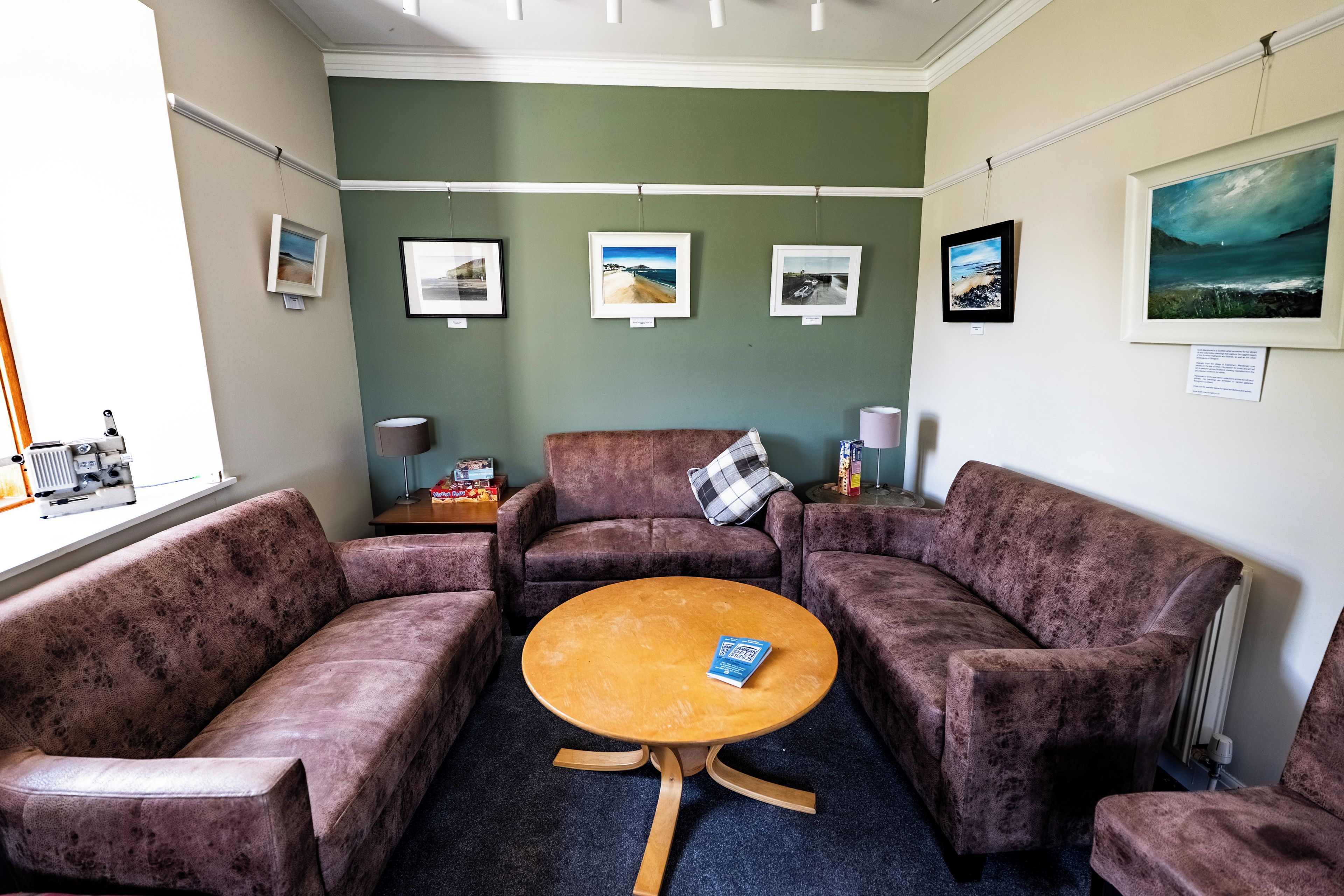 A room with a round wooden table, four cushioned chairs, a small side table with a lamp, and framed pictures on the wall.