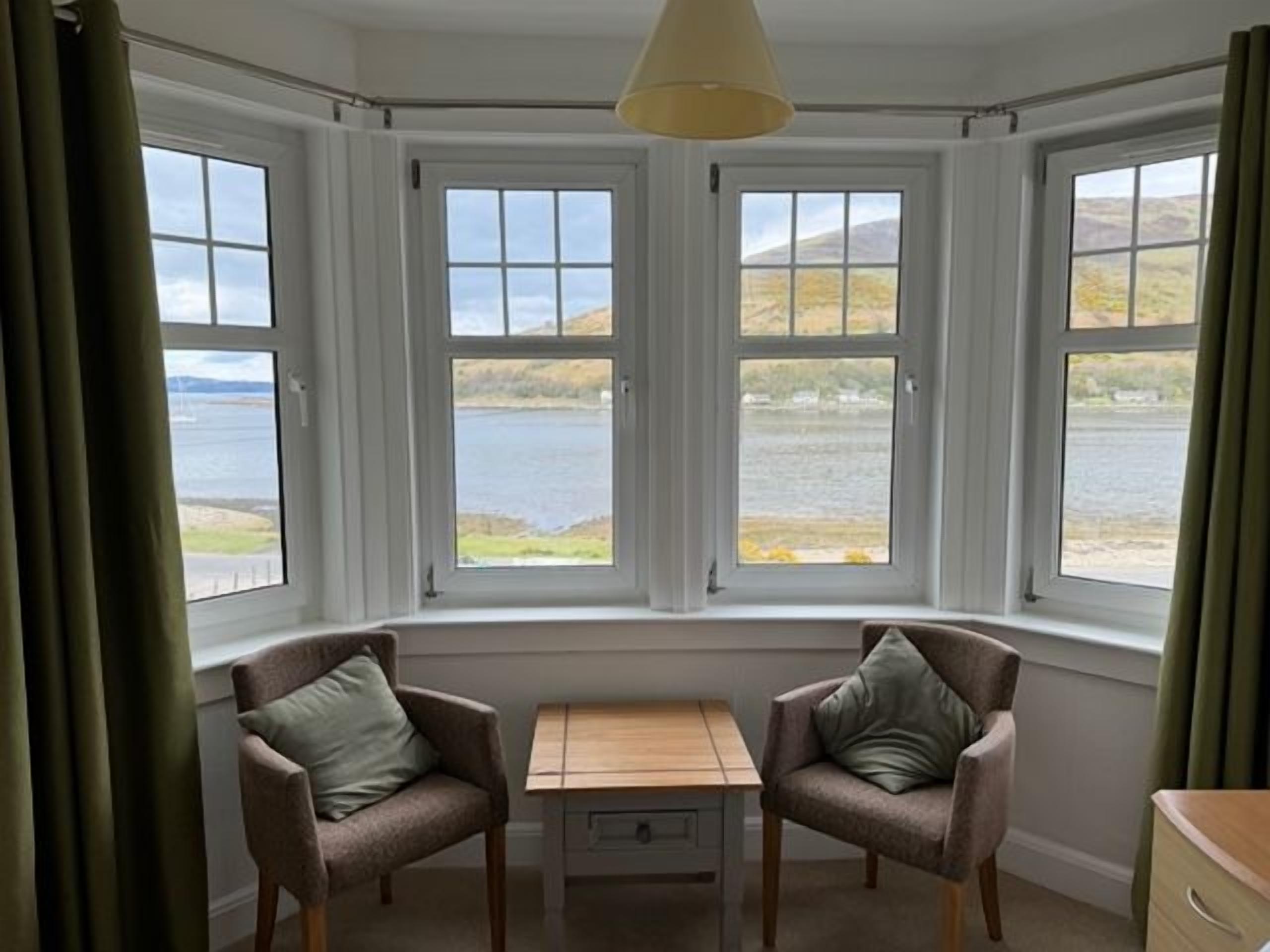 A room with a bay window overlooking a body of water, two armchairs, a small table, and a lamp.