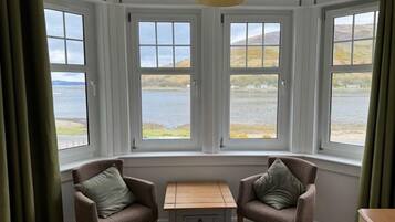 A room with a bay window overlooking a body of water, two armchairs, a small table, and a lamp.