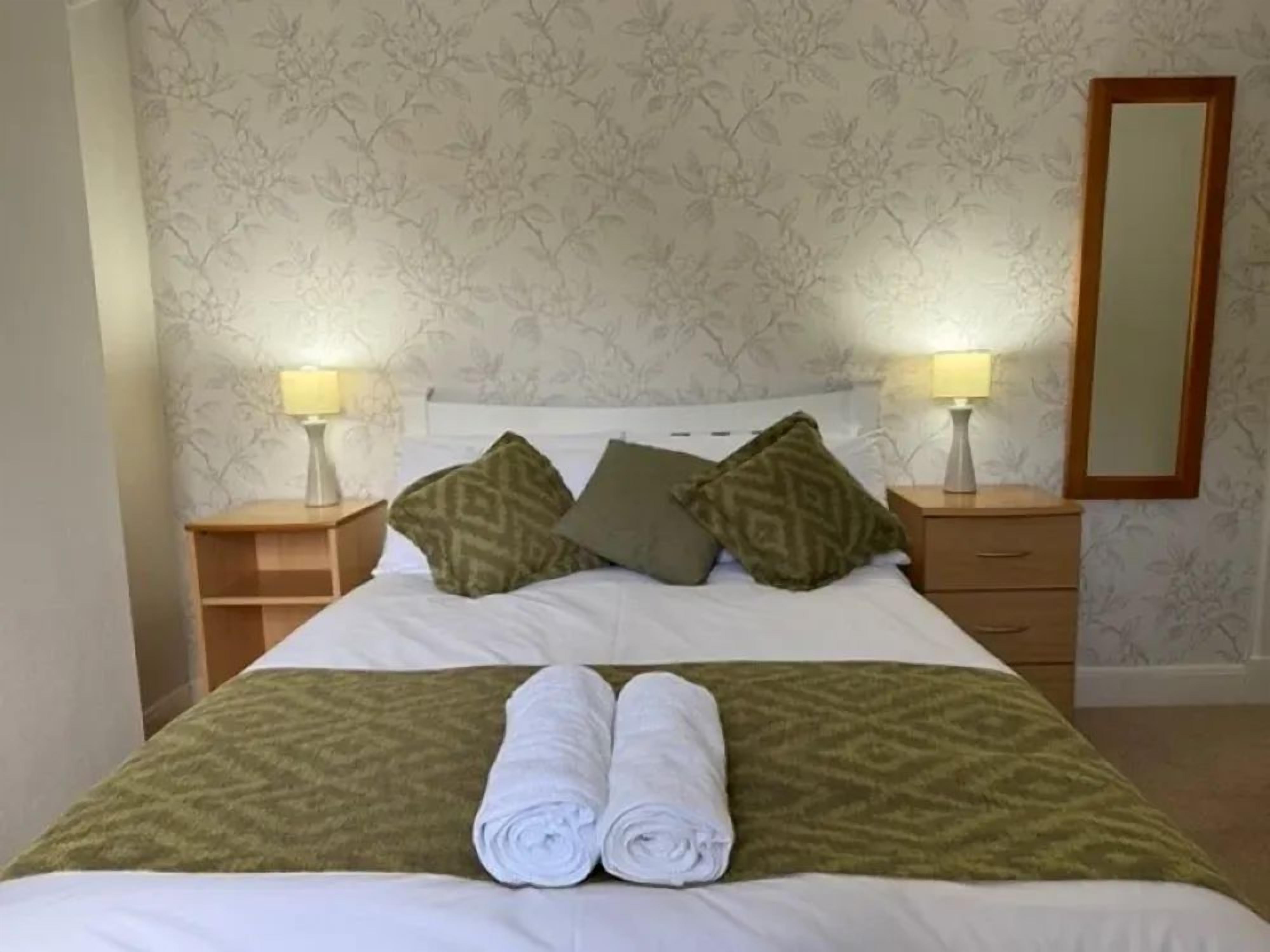Cama bem arrumada com travesseiros e toalhas dobradas, mesa de cabeceira com abajur e espelho na parede.