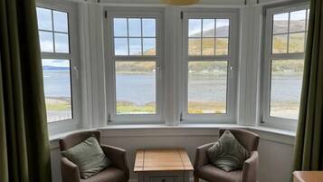 A room with a bay window overlooking a lake, two armchairs, a small table, and a hanging light fixture.