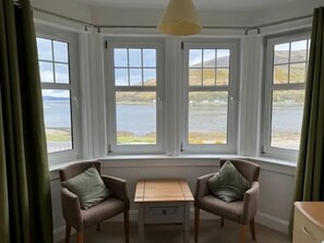 A room with a bay window overlooking a lake, two armchairs, a small table, and a hanging light fixture.