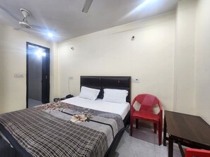 A bedroom with a bed, a chair, a table, a ceiling fan, and a door. - Oly Hotel Star Inn (New Delhi)