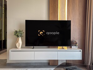 Ein Flachbildfernseher auf einer weißen Konsole mit einem Logo auf dem Bildschirm, eine Vase mit Grünpflanzen und eine Fernbedienung auf der Konsole.