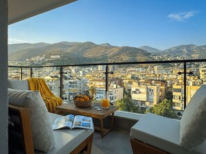 A balcony with a view of a cityscape and mountains. There is a table with a basket of fruit and a glass of orange juice. - Qoople Prive 42 - Cozy 1-bedroom apartment in Alanya (Alanya)