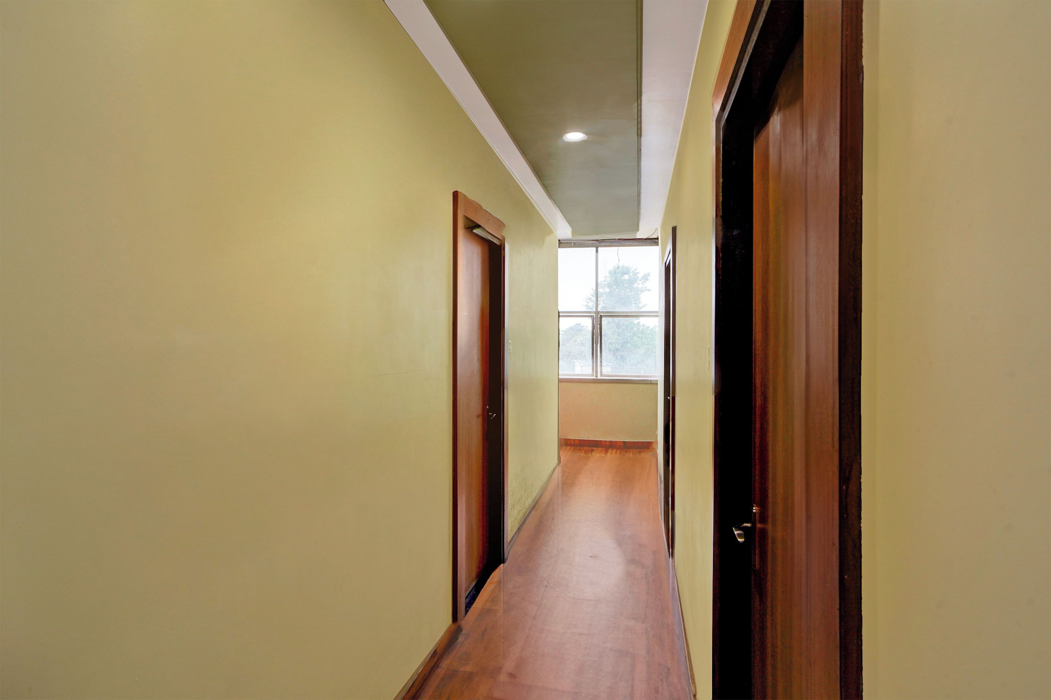 A narrow hallway with wooden flooring and doors on both sides, leading to a room with a window.