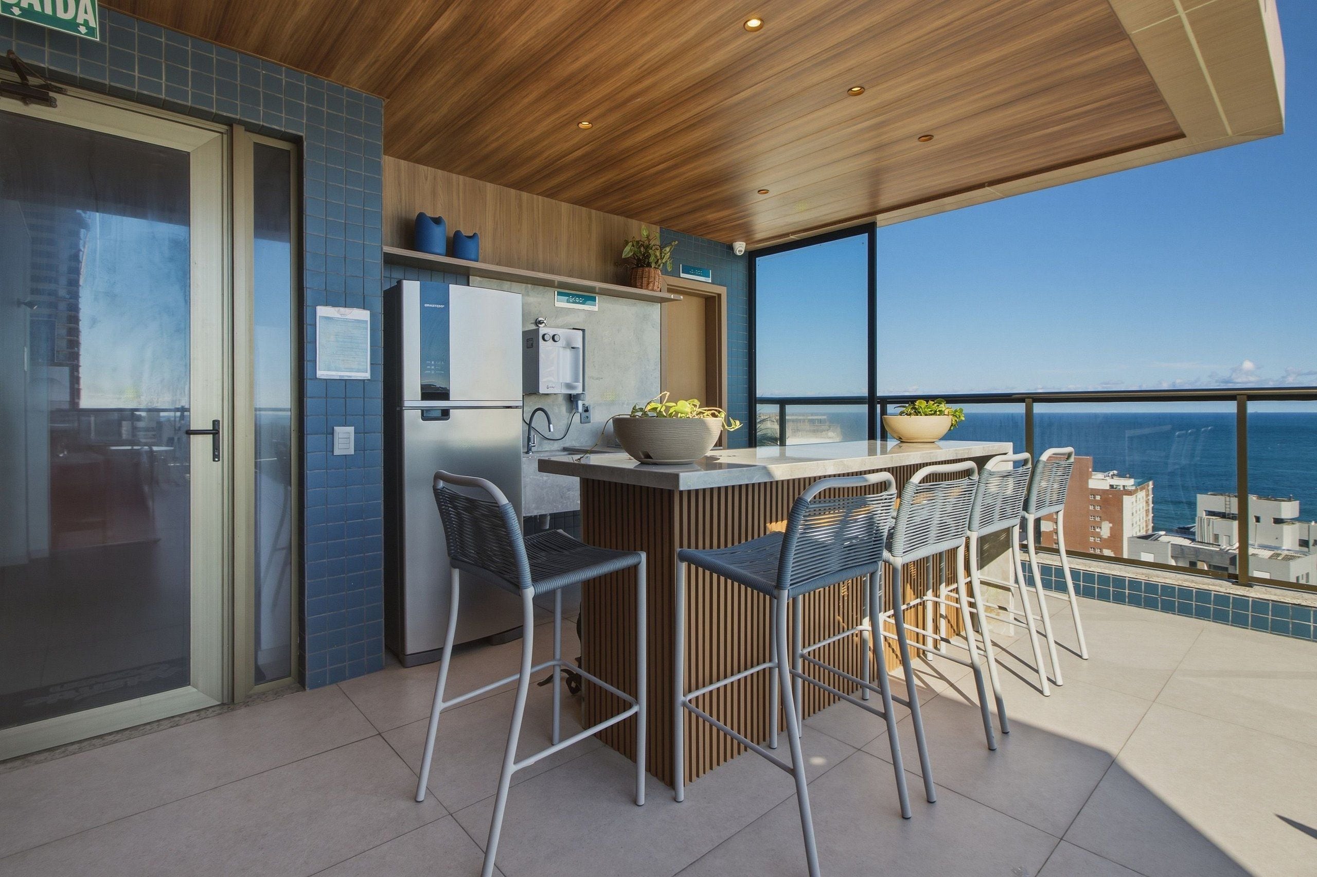 A balcony with a view of the sea, a bar area with chairs, a refrigerator, and a coffee machine.