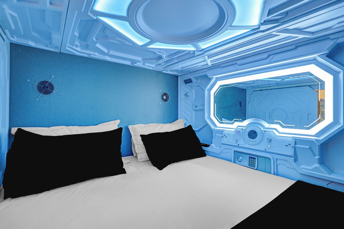 A futuristic room with a bed, a control panel, and a large window.