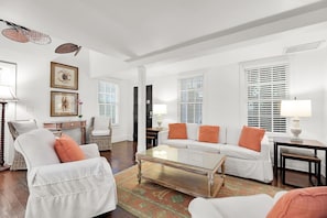 A living room with a white sofa, a coffee table, and a wicker chair. There are two lamps on side tables, and a rug on the floor. - Jacaranda Cottage 18+ Adult Only cottage near Duval St (Key West)