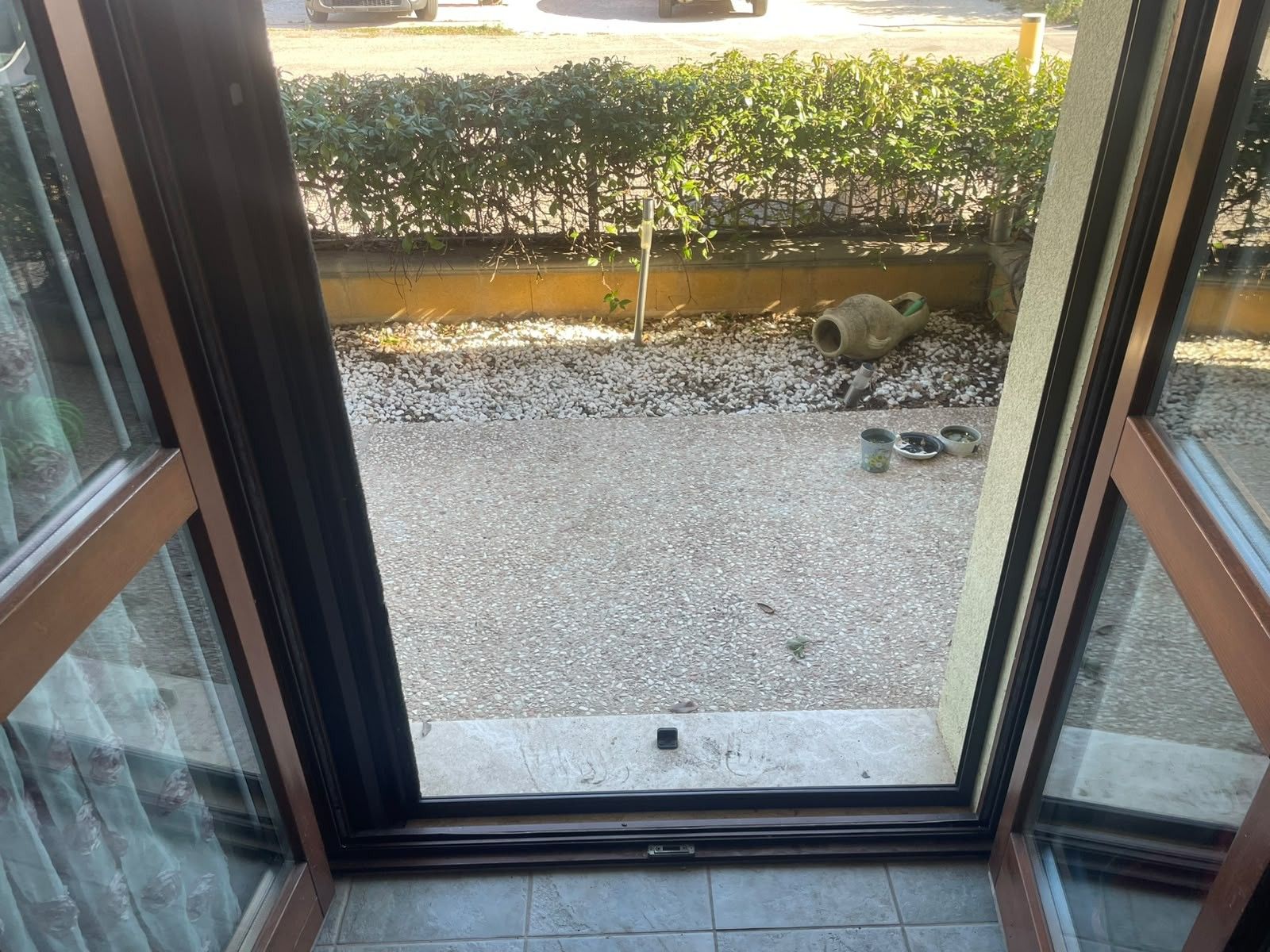 Visão através de uma porta de vidro mostrando uma área com cascalho, uma pequena planta, um recipiente de metal e um objeto amarelo.
