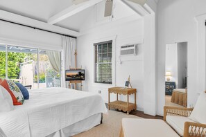 A bedroom with a bed, a television on a stand, a wicker side table, a chair, and a window with blinds. - Bougainvillea Manor 18+Adult Only retreat near Duval St (Key West)