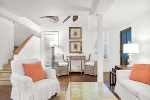 A living room with a white sofa, a coffee table, wicker chairs, a small table with a lamp, and artwork on the wall. - Orchid Cottage, Two cottages, one 18+ Adult Only garden retreat (Key West)