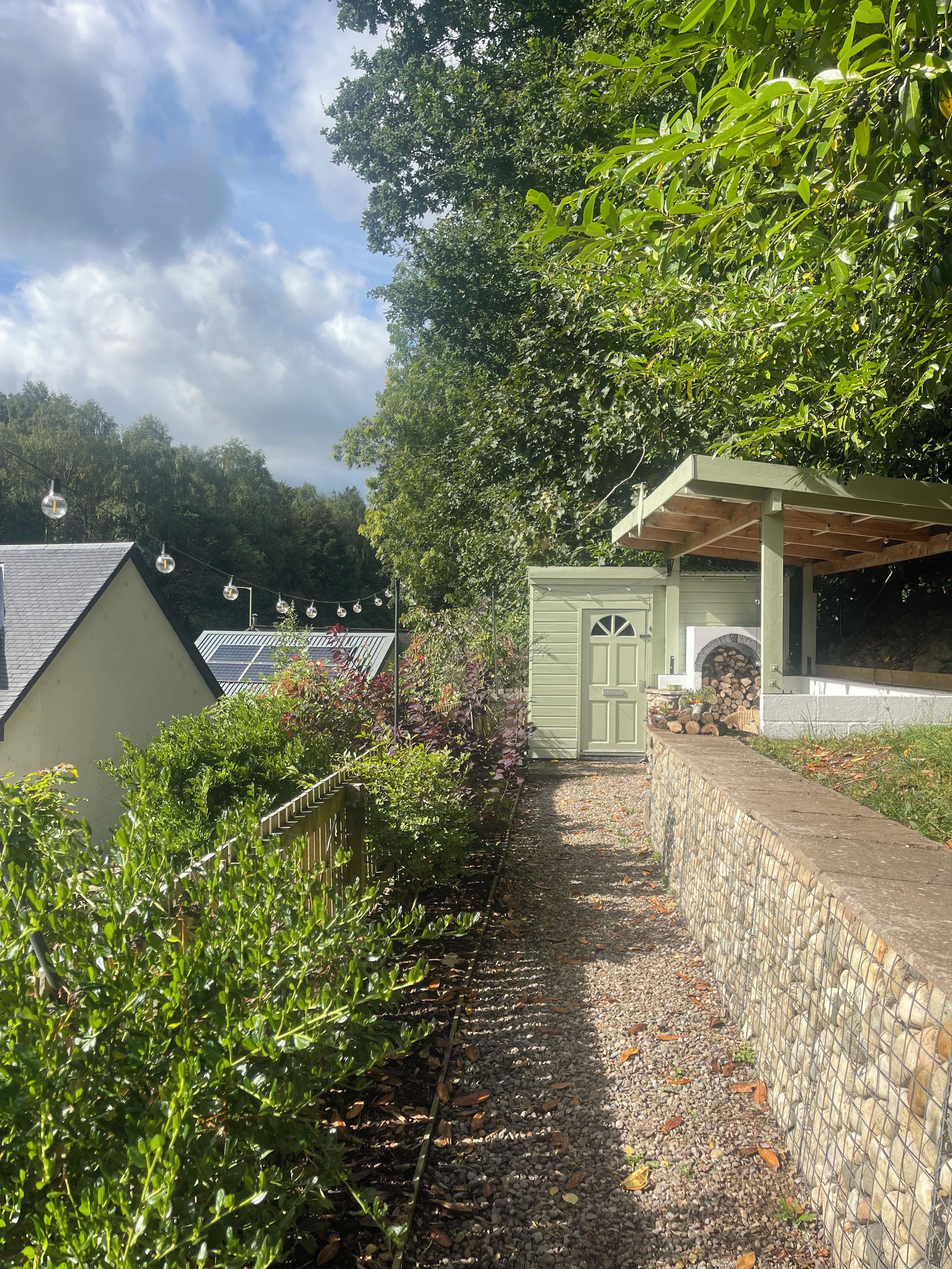 A pathway leading to a green structure with a door, surrounded by greenery and a stone wall.