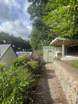 A pathway leading to a green structure with a door, surrounded by greenery and a stone wall.