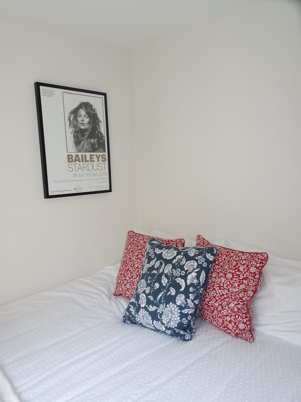 A bedroom with a bed, white bedding, and three pillows with floral patterns. A framed poster is on the wall.