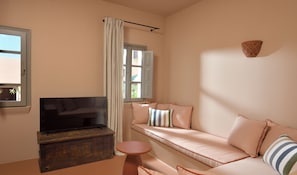 A living room with a sofa, a television on a wooden cabinet, a small round table, and a window with curtains. - Terra Verde Oia Suites - Patio Grand Suite with Inner Courtyard View (Oia)