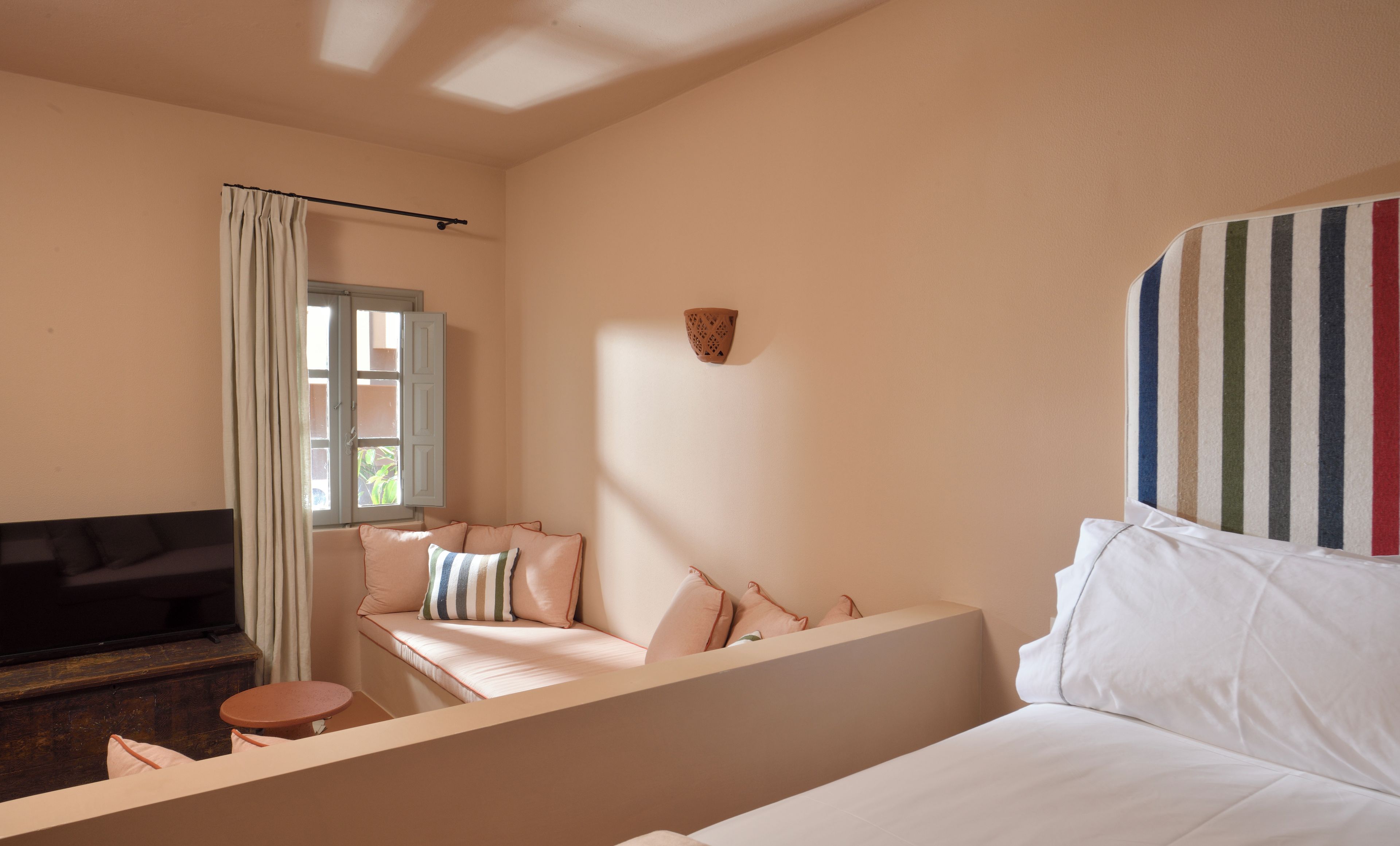 A bedroom with a bed, a sofa, a small table, a window with curtains, and a wall-mounted light fixture.