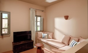 A living room with a sofa, a TV, a small table, and a window with curtains. - Terra Verde Oia Suites - Patio Grand Suite with Inner Courtyard View (Oia)