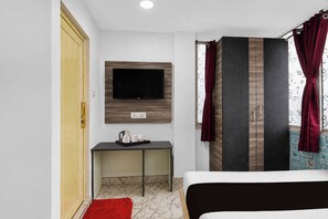 Una habitación de hotel con una cama, un televisor montado en la pared, un escritorio pequeño con una tetera y tazas, y una alfombra roja.