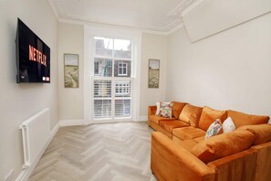Un salón con un televisor de pantalla plana grande montado en la pared, un sofá con cojines, un radiador y una ventana con vista a un edificio.
