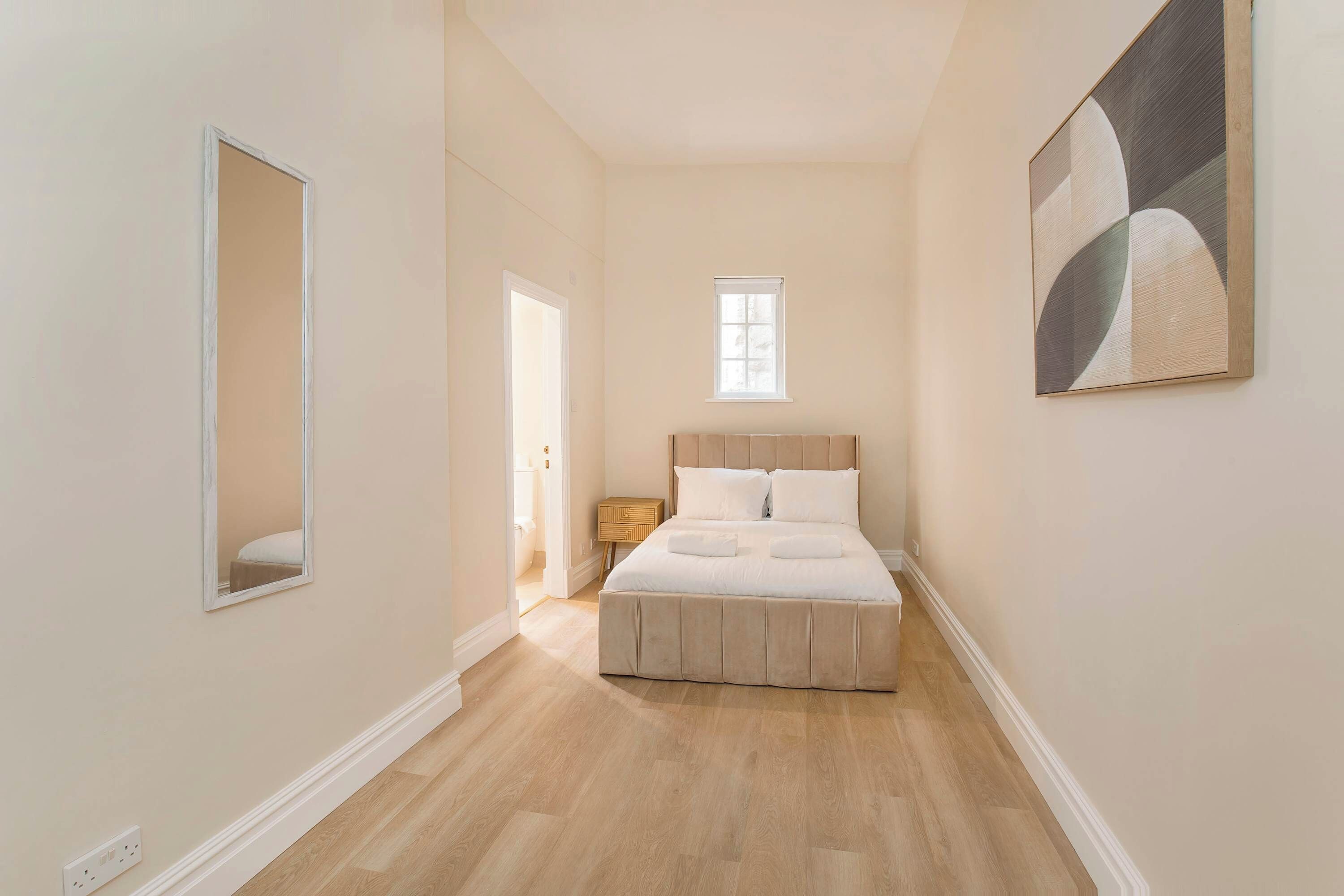Un dormitorio con una cama, una mesita de noche, un espejo, una ventana y un cuadro en la pared.