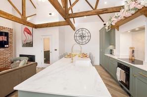 A modern kitchen with a central island, wooden beams, a large clock, and a TV mounted on a brick wall. - Ousegate Skyline Retreat, sleeps 6 (York)