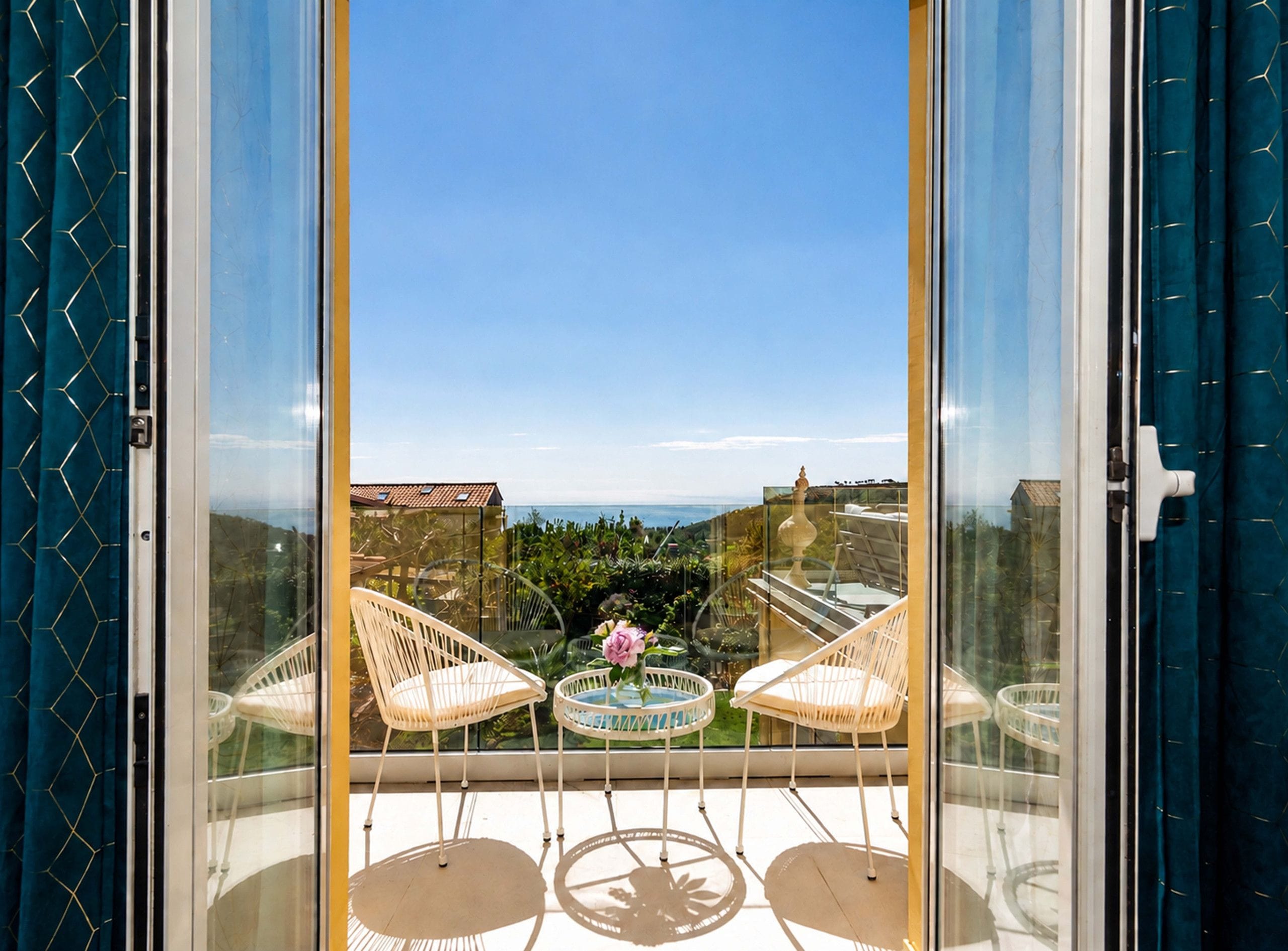A balcony with two white wicker chairs and a small round table, overlooking a garden and a distant view.