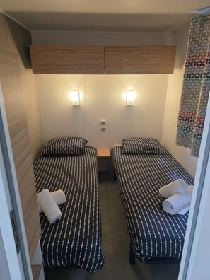 A small, compact room with two single beds, each with a pillow and a rolled towel. There is a small table between the beds, and the room has a simple, functional design with a light fixture above the table. - 5-Star Houlgate Campground - 2-bedroom, 4-person mobile home with AC, facing the pool. 1.5 km from the beach (houlgate)