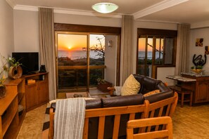 A living room with a sofa, a television, and a view of the sunset through large windows.