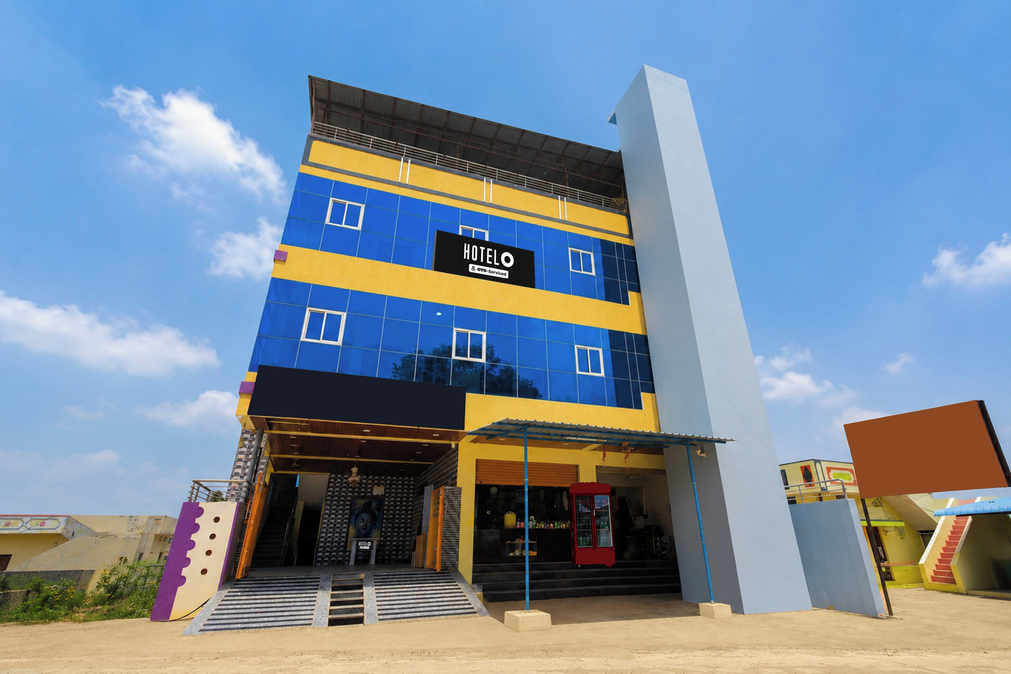 A multi-story hotel with a blue and yellow facade, a visible entrance with steps, and a sign indicating it is serviced by OYO.