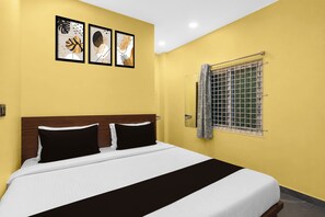 A hotel room with a bed, a window with a metal grill, and three framed artworks on the wall.