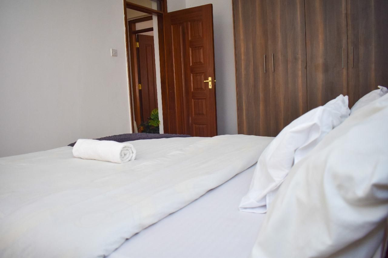 Een net opgemaakt bed met een witte dekbedovertrek en een opgerolde witte handdoek, een houten kledingkast en een gedeeltelijk open deur naar een andere kamer.