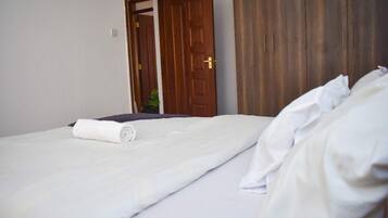 Cama bem arrumada com edredom branco e toalha branca enrolada, guarda-roupa de madeira e porta entreaberta para outro quarto.