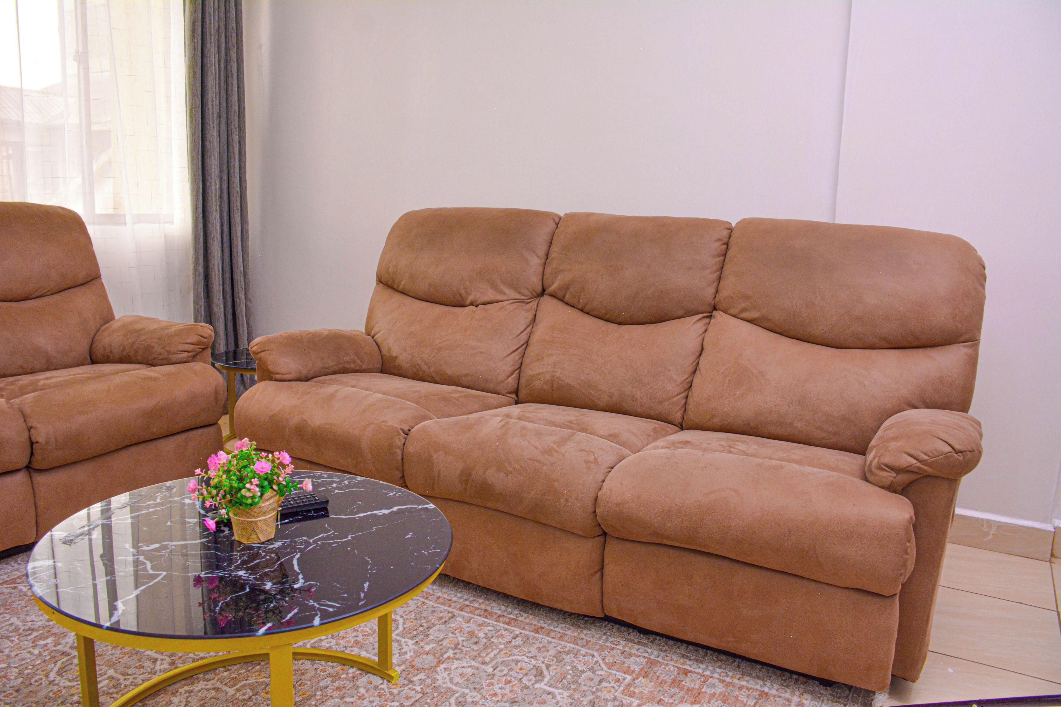 Una sala de estar con un sofá marrón, una mesa de centro de vidrio con un jarrón de flores y una alfombra estampada.
