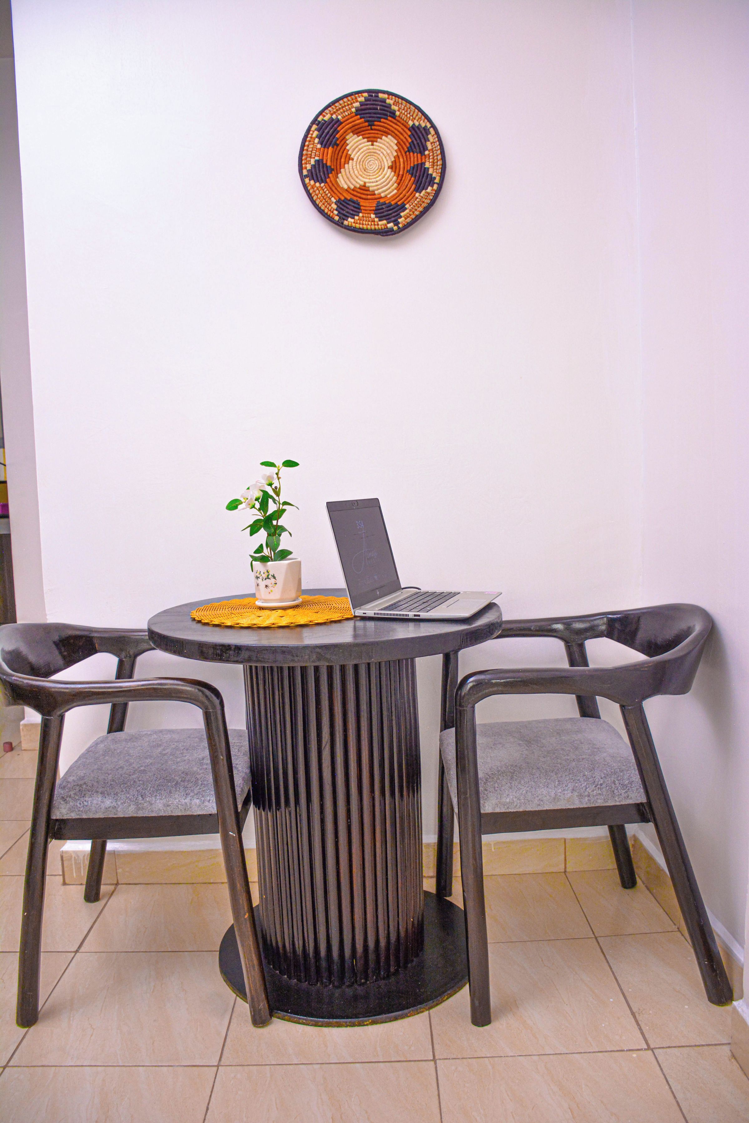 Una mesa redonda con una computadora portátil y una planta en maceta, acompañada por dos sillas, frente a una pared lisa con un plato decorativo arriba.