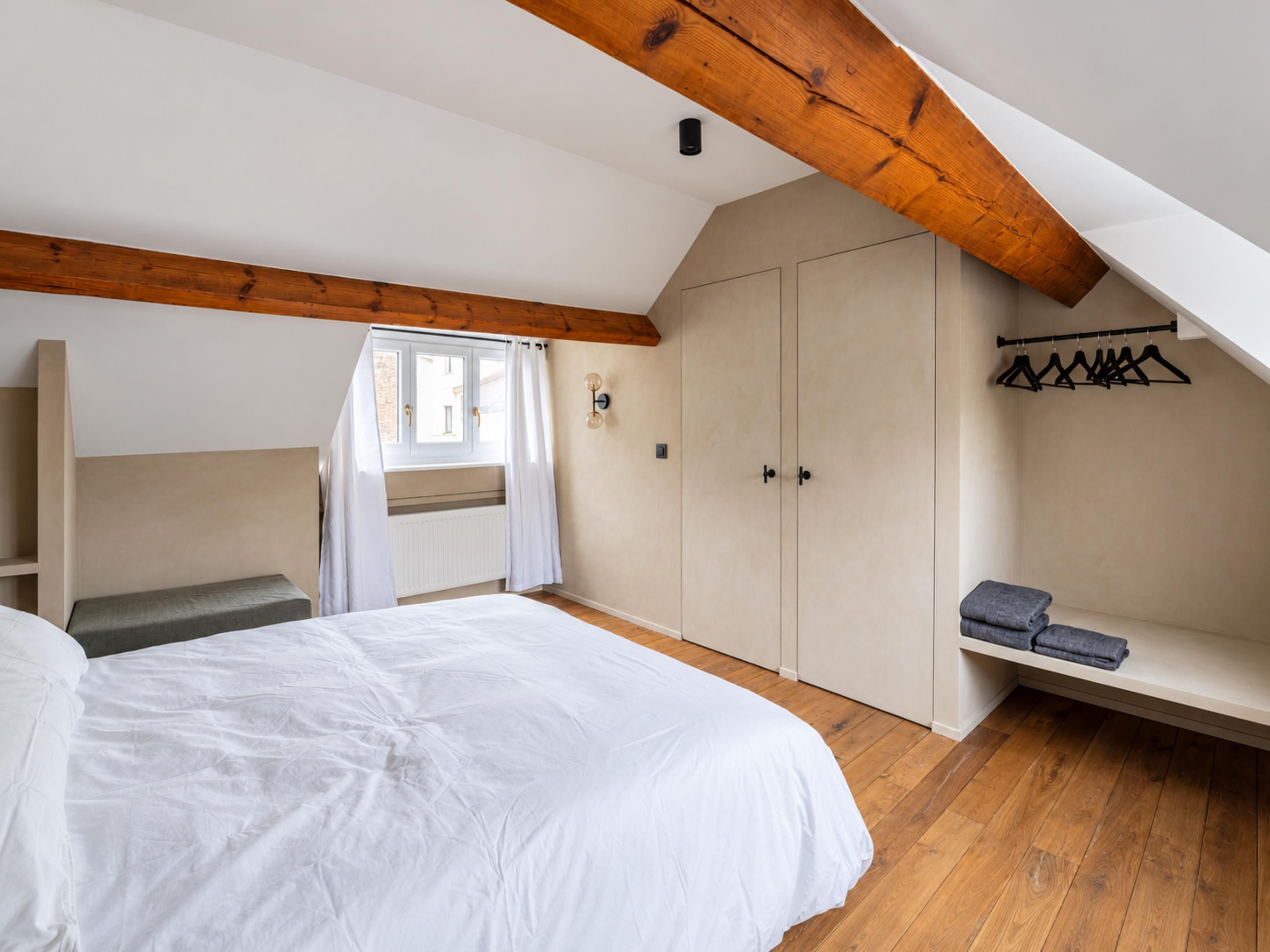 A bedroom with a large bed, a wardrobe, a bench, and a window with curtains.