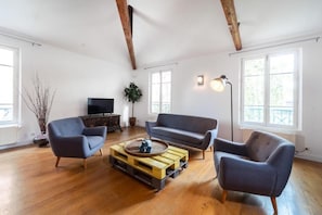 A spacious living room with wooden floors, a grey sofa, a blue armchair, and a wooden coffee table.