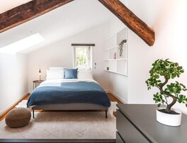 Un dormitorio con techo inclinado, una cama con manta azul, una planta en maceta y un estante blanco.