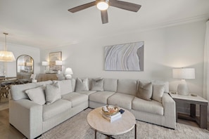 A modern living room with a sectional sofa, a round coffee table, and a ceiling fan. - Romar House 1004A by Liquid Life (Orange Beach)