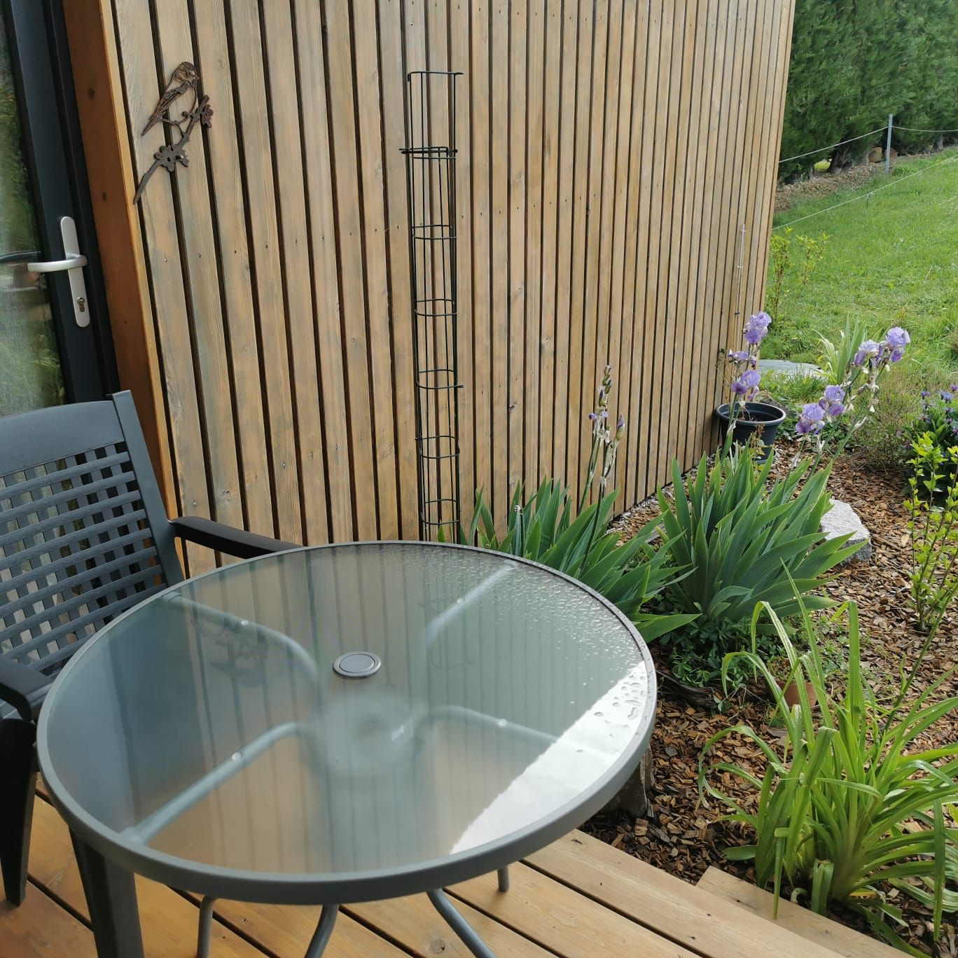 Een tafel met glazen blad en metalen poten, een zwarte stoel en een houten terras.