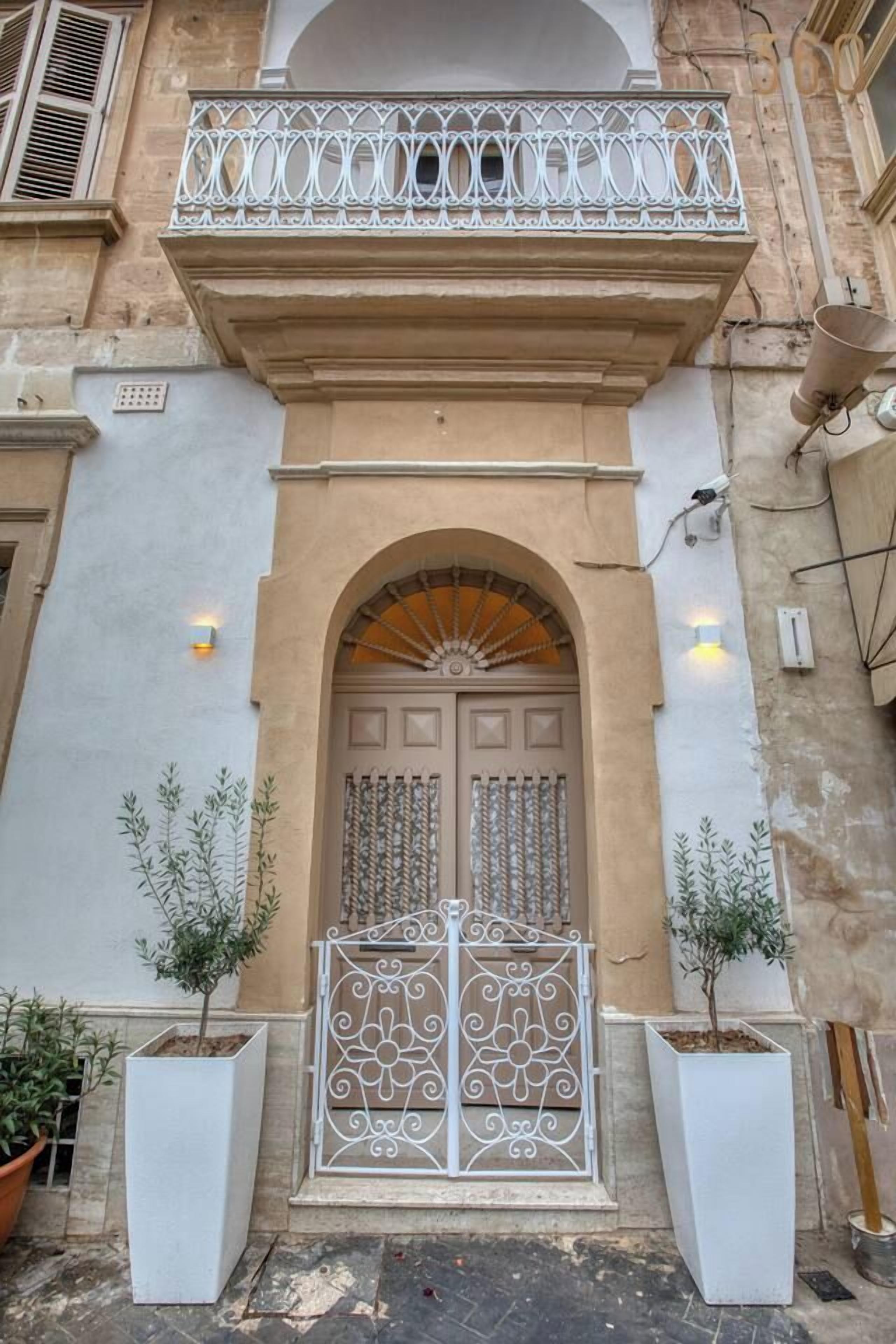 A building facade with a central arched entrance, a decorative balcony, and two potted plants.