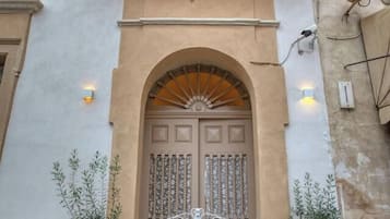 A building facade with a central arched entrance, a decorative balcony, and two potted plants.