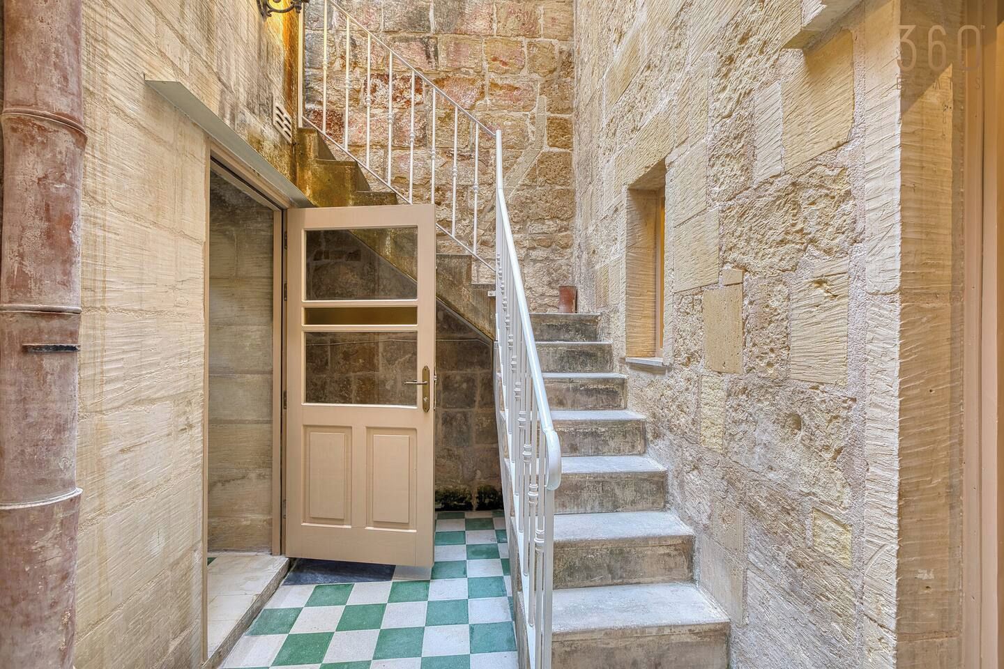 Un estrecho pasillo con piso a cuadros, una puerta de madera y una escalera con paredes de piedra.