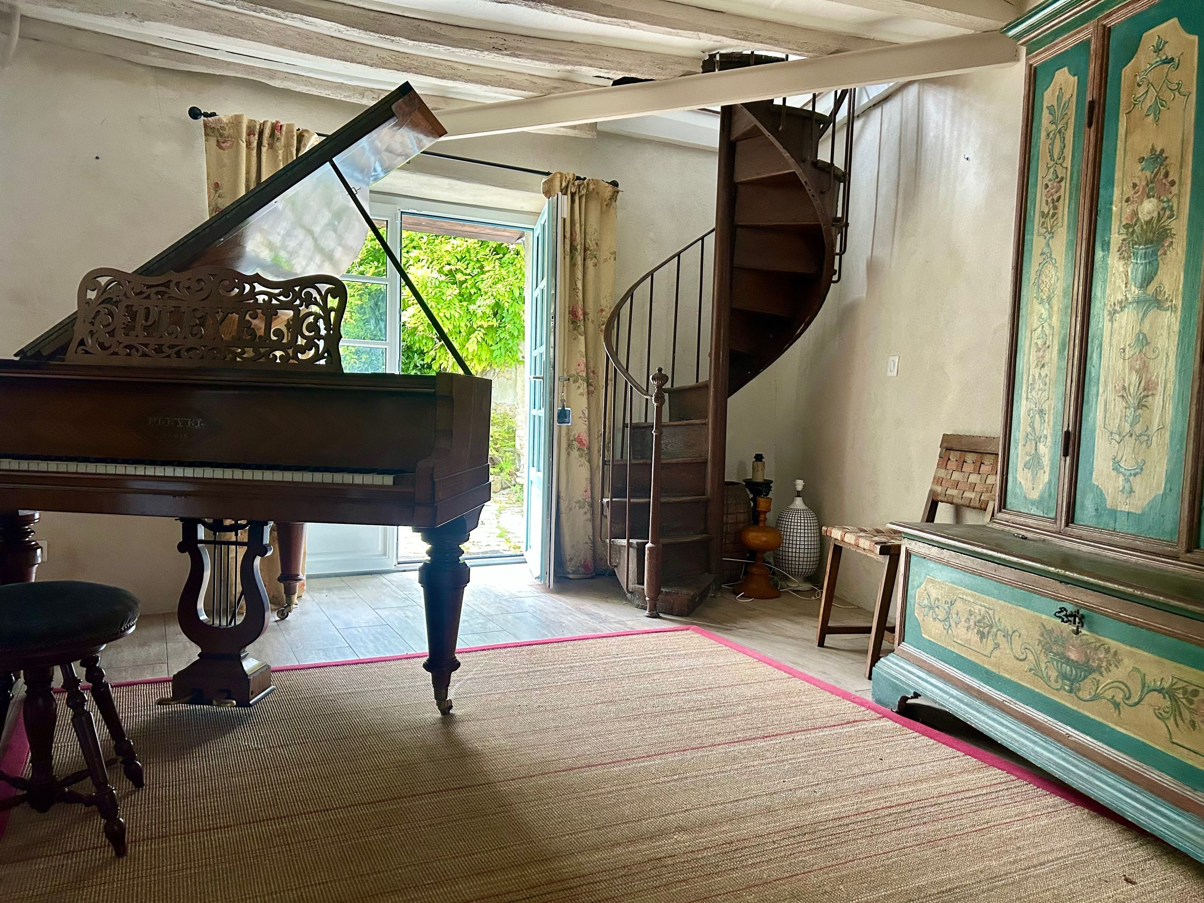 Un piano de cola en una habitación con una escalera, un mueble grande con diseños florales y vistas al exterior con follaje.