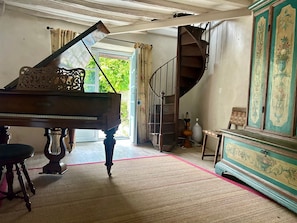 Un piano de cola en una habitación con una escalera, un mueble grande con diseños florales y vistas al exterior con follaje.