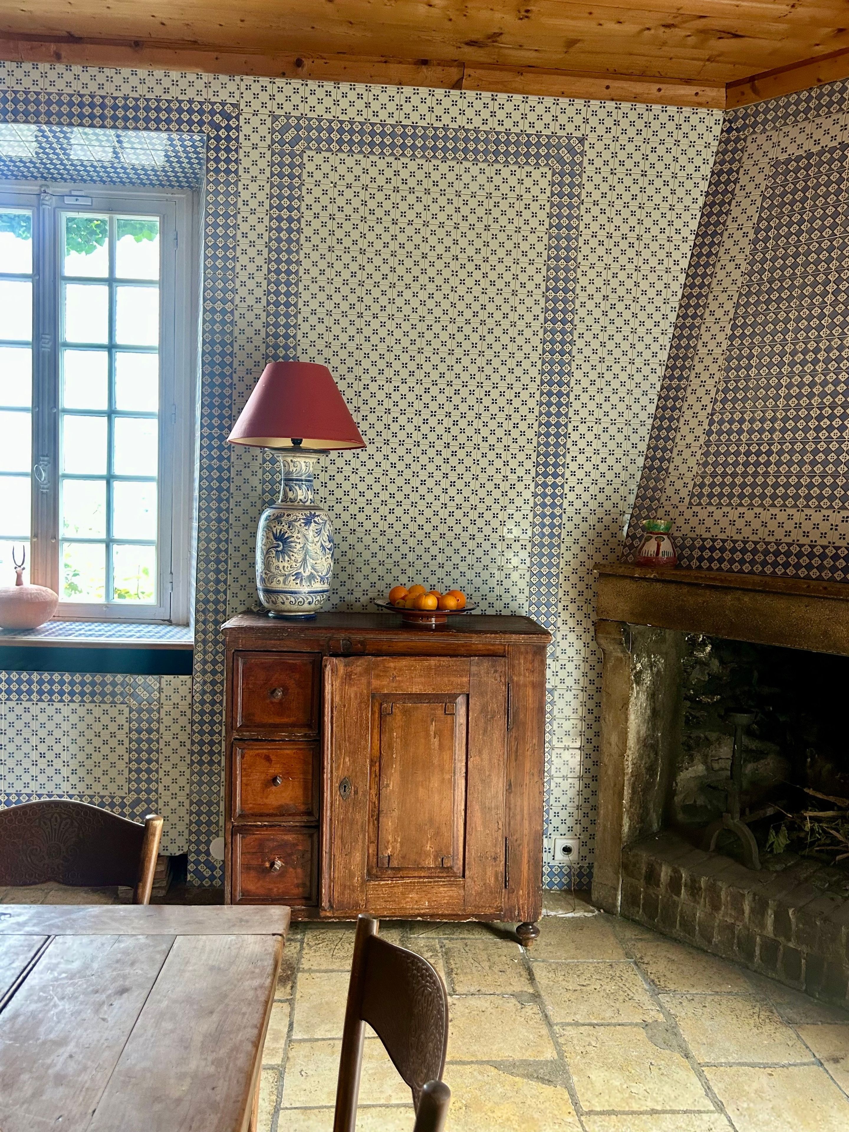 Ein Zimmer mit ornamentalem Wandpapier, einem Holzschrank, einem Tisch und einem Kamin.