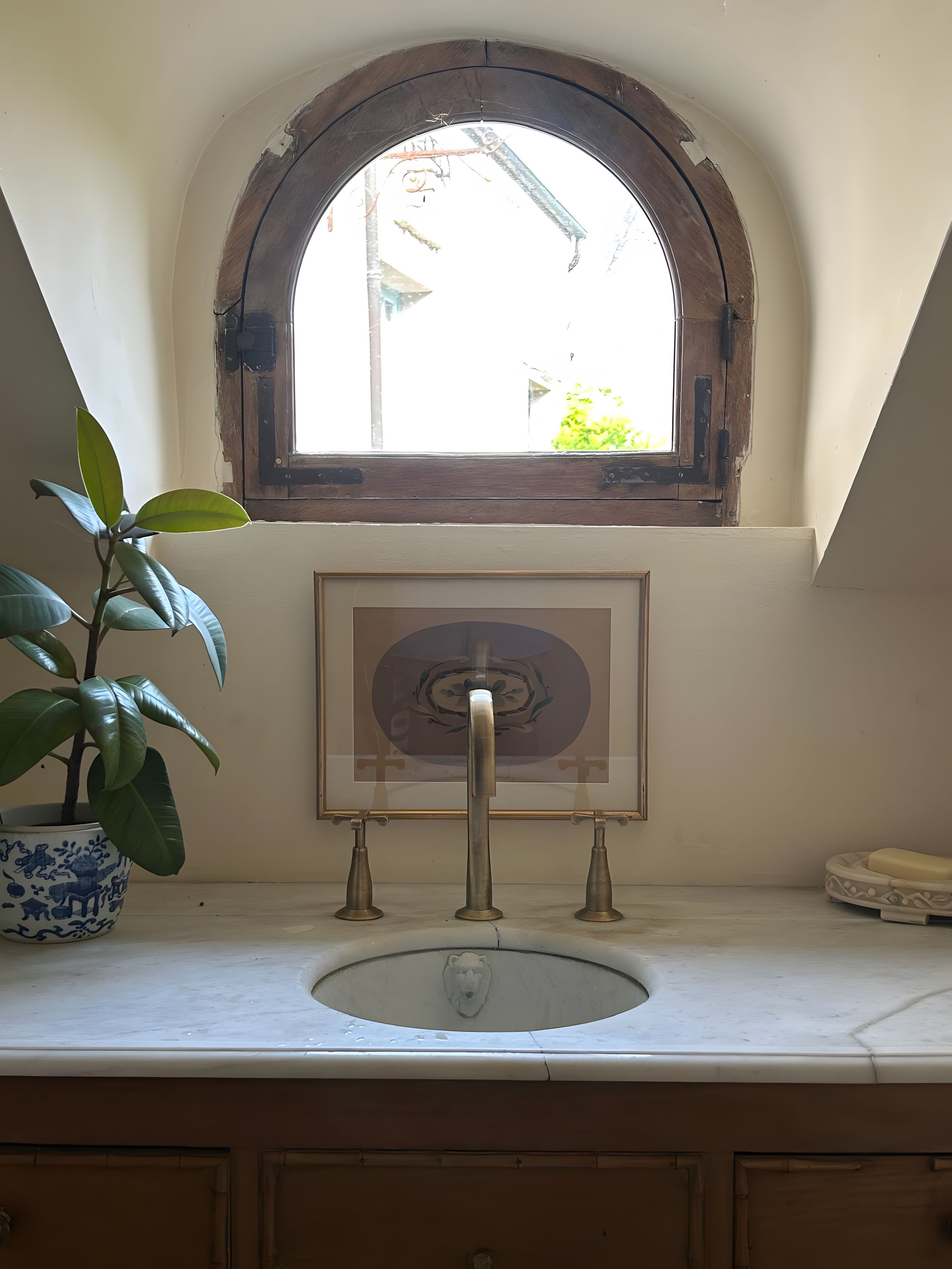 Une salle de bain avec un lavabo, une plante en pot, un tableau encadré et une fenêtre donnant sur un paysage verdoyant.