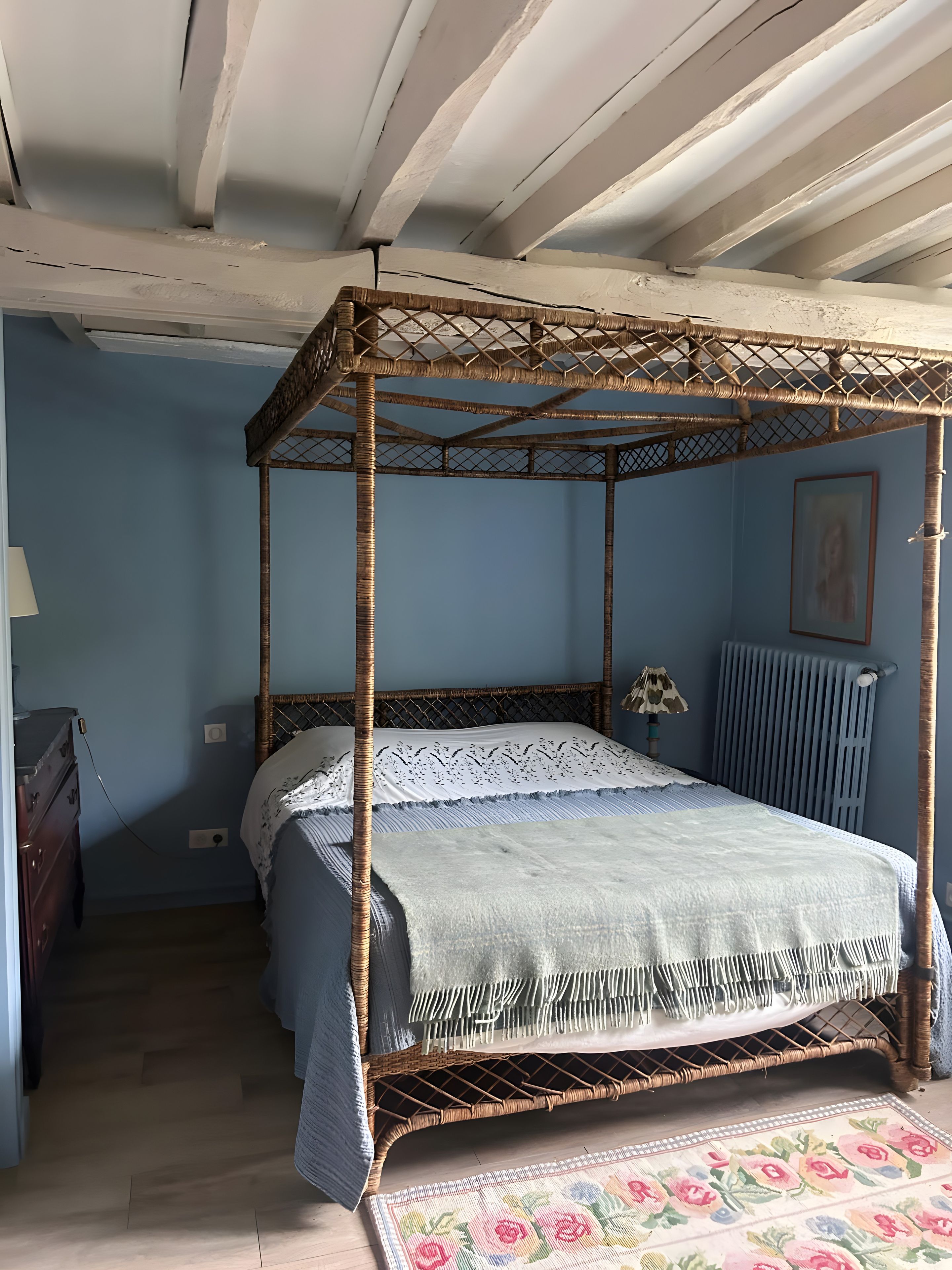 A four-poster bed with a woven headboard, a patterned bedspread, and a floral rug on a wooden floor.