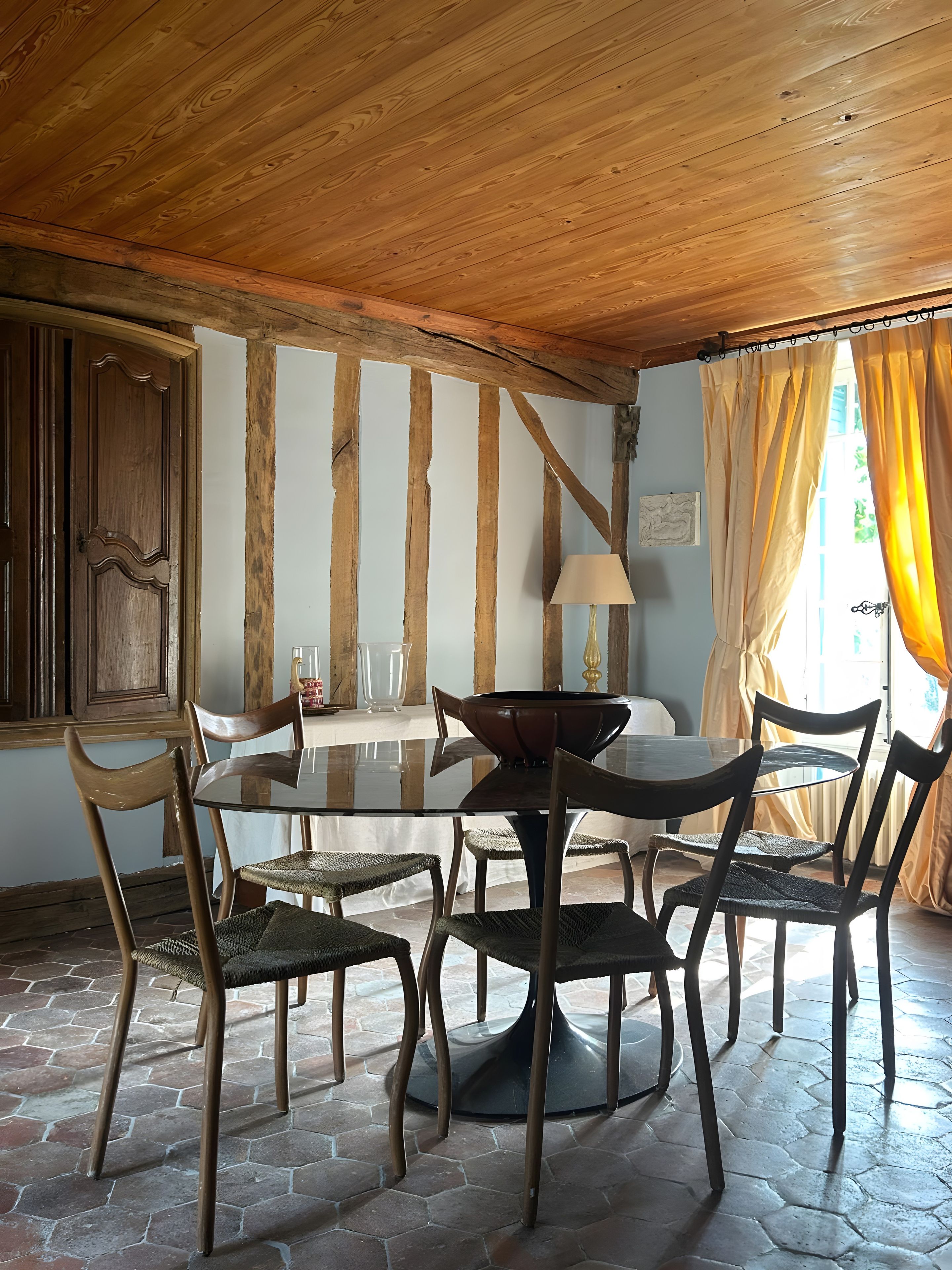 Een eetgedeelte met een ronde glazen tafel, zes houten stoelen, een houten plafond en een raam met gordijnen.